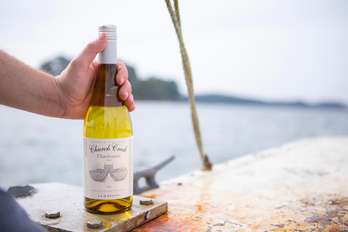 A hand holds a bottle of Chatham Vineyards at Church Creek Chardonnay on a boat. The label features a vineyard illustration and text. The boat's deck and a mooring cleat are visible, with an out-of-focus body of water and shoreline in the background.