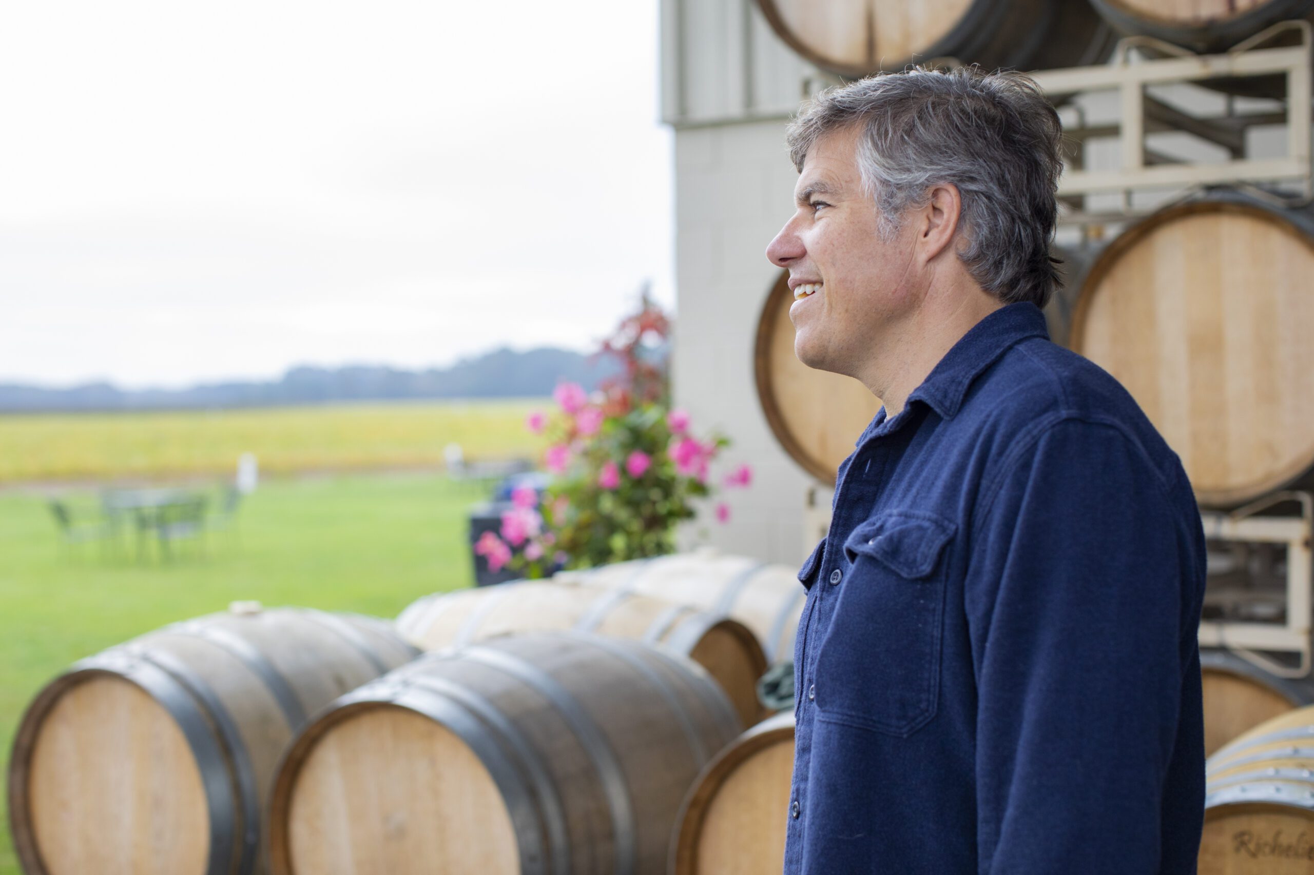 A man in a blue shirt stands next to wine barrels while looking over Chatham Vineyards