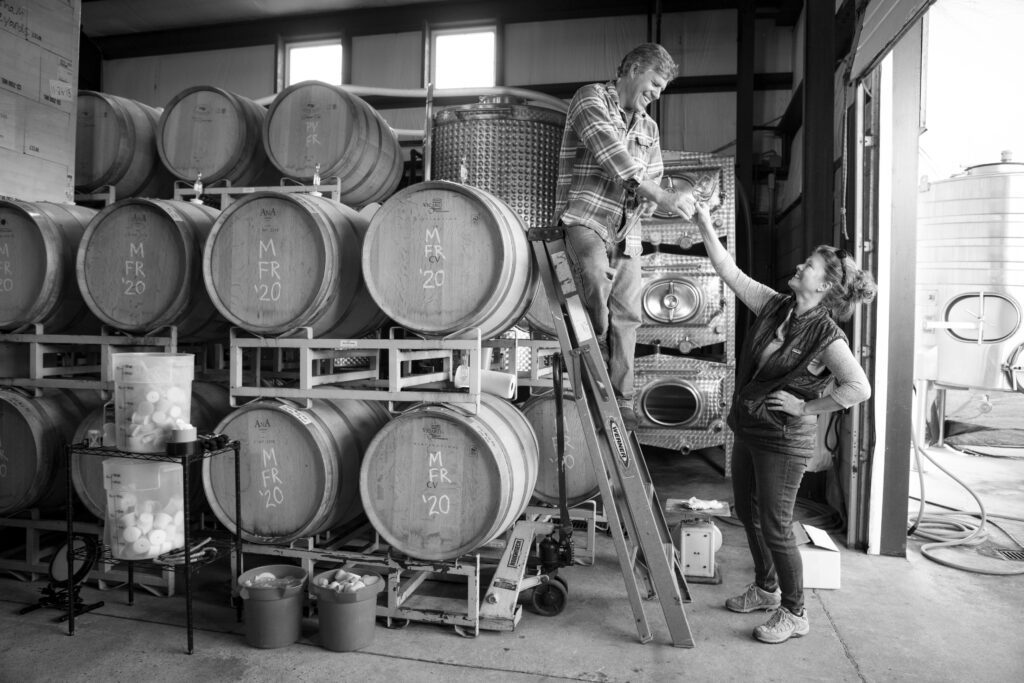 A man standing on a ladder in front of barrels of wine at Chatham Vineyards hands a glass of wine to a woman