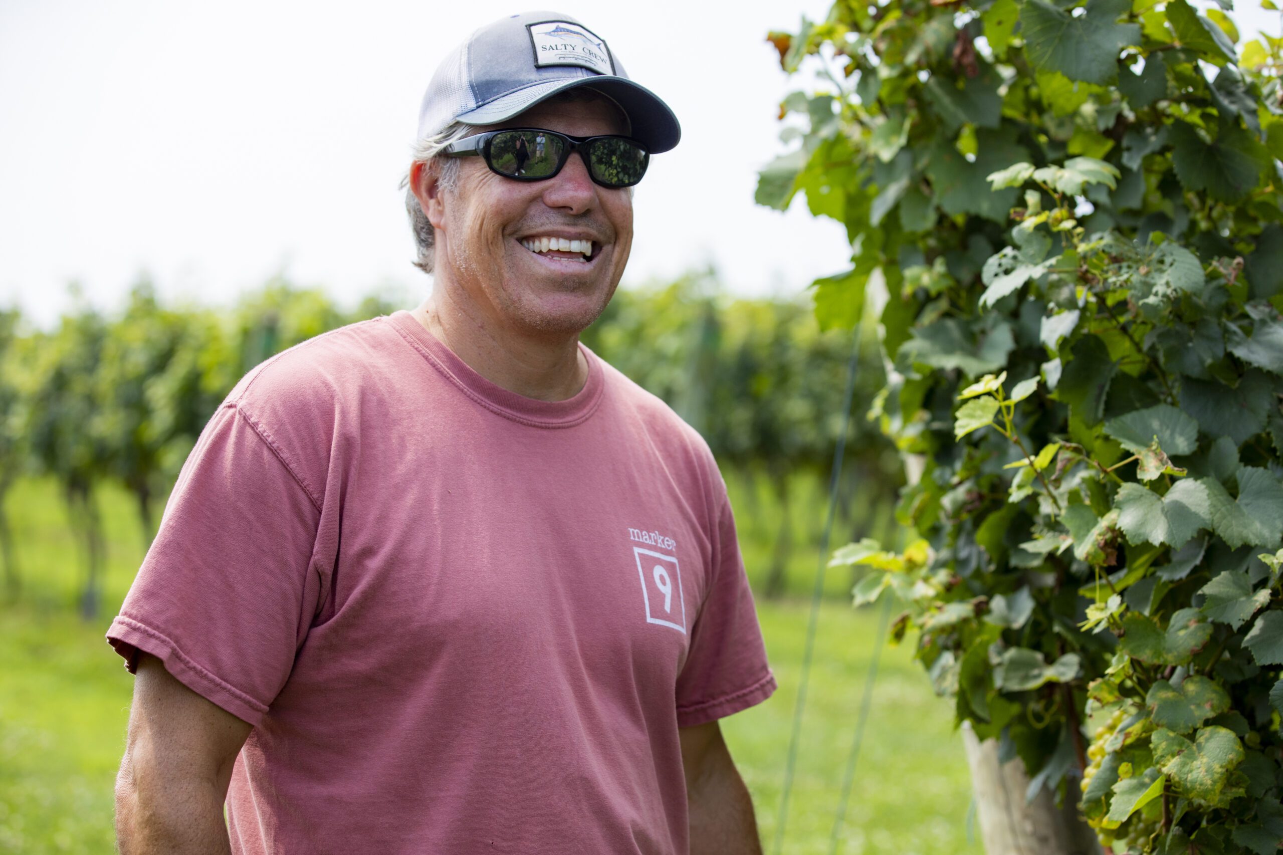 A man wearing a baseball hat and a red shirt smiles in the vineyard at Chatham Vineyards