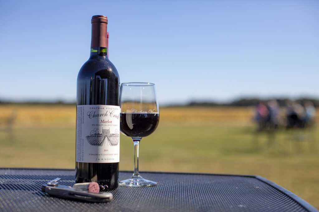 A bottle of red wine and a glass of wine sit on a black metal table outdoors. The bottle is labeled "Cabernet Franc Merlot." A wine cork, corkscrew, and bottle cap are also seen on the table. In the background, there is a blurred view of a grassy field and people sitting.