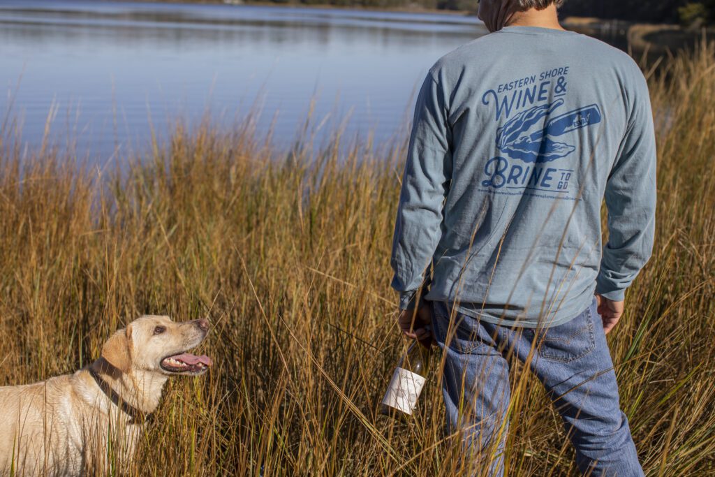 man staring out at the water wearing a Wine and brine shirt and holding a bottle of Chatham wine with a yellow lab next to him