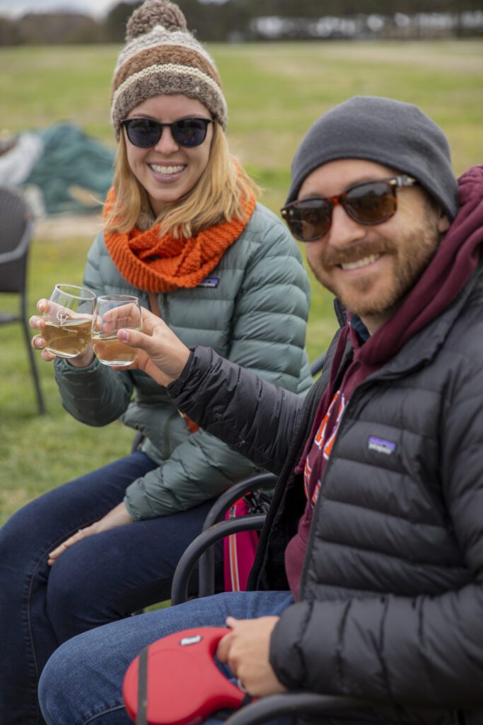 couple enjoying glasses of white wine in winter coats outside while smiling at the camera