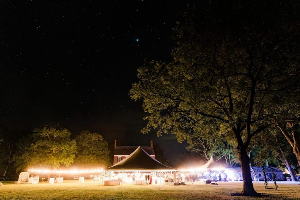 A nighttime outdoor wedding event with a large tent lit by string lights on a lawn, surrounded by trees. A house is visible in the background under a starry sky.