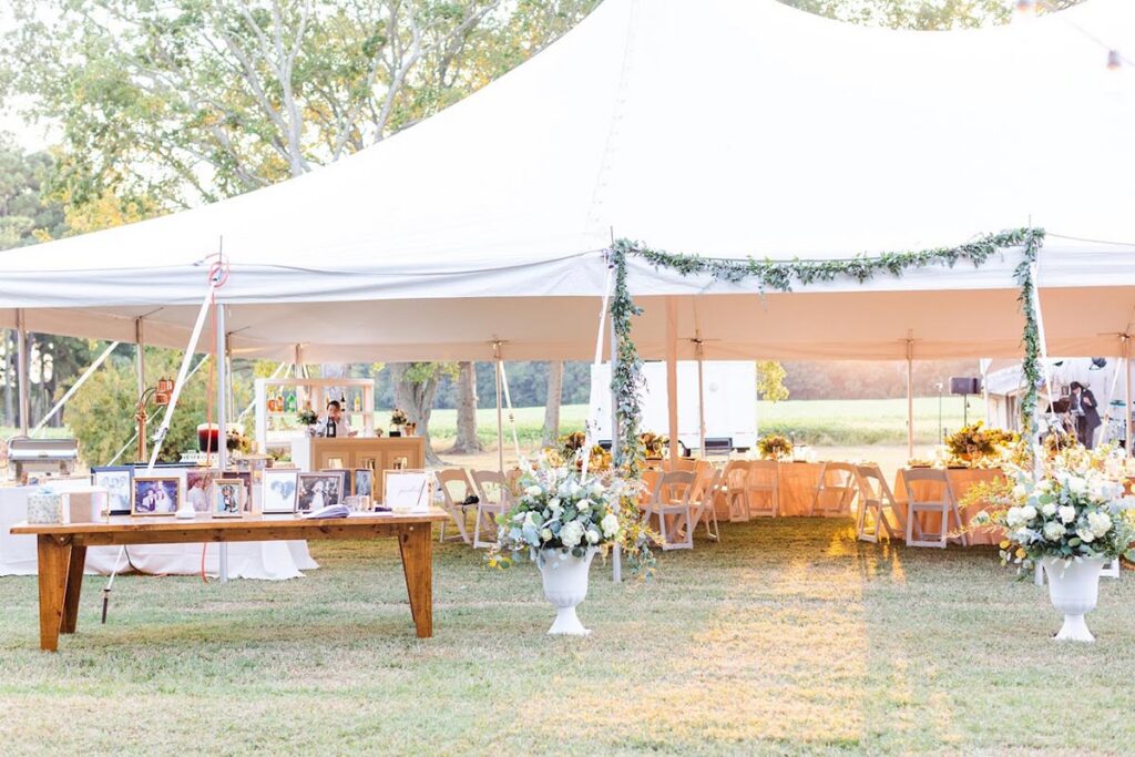 Outdoor wedding tent decorated with greenery and flowers, featuring white chairs and tables inside. A wooden table with framed photos and two large floral arrangements stand on the grass outside the tent.