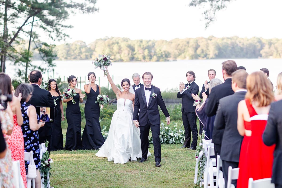 A bride and groom walk down the aisle outdoors, smiling and holding hands, as guests applaud. Bridesmaids in black dresses and groomsmen in black suits stand nearby. A lake and trees are in the background.